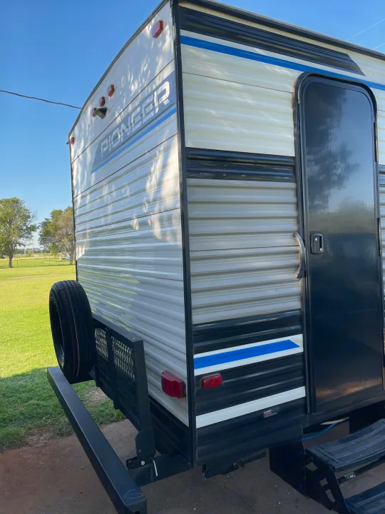 Exterior of a white and gray travel trailer, an rv rental, with a spare tire and utility rack, parked in a grassy field.. Heartland RVs Pioneer 2023