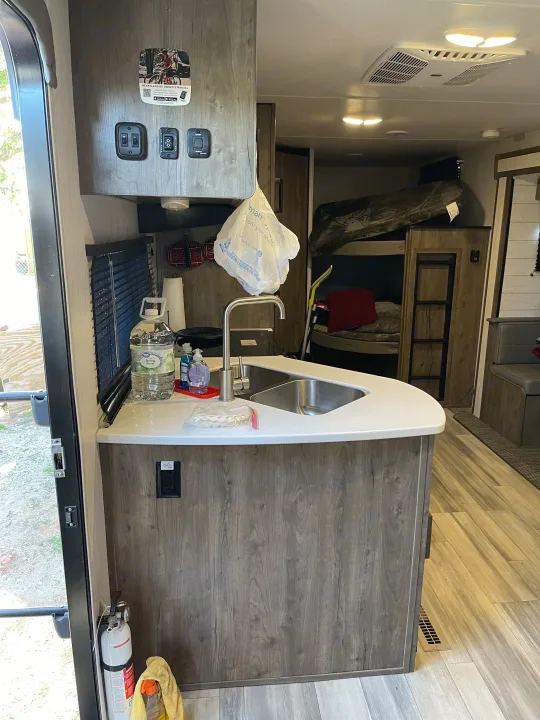 Inside an RV rental, a modern kitchen with a sink and controls is featured, with bunk beds and seating visible in the background.. Heartland RVs Pioneer 2023