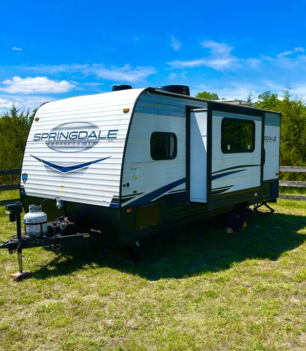 A white Springdale Keystone RV mini, a compact travel trailer for rv rental, sits in a sunny grassy field.. Keystone RV Springdale 2023