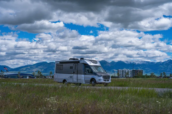 A sleek rv rental parked on a path, surrounded by green fields, buildings, and snow-capped mountains under a dramatic cloudy sky.. Regency RV Ultra Brougham 2021