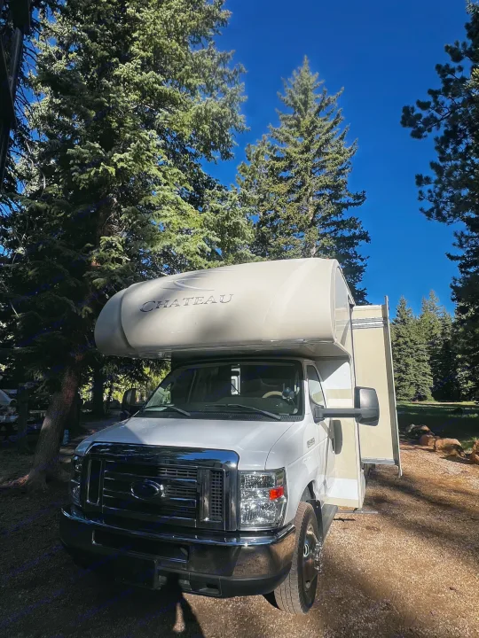 A white Chateau RV rental rests among tall evergreen trees under a bright blue sky, ideal for outdoor travel and camping.. Thor Chateau 2017