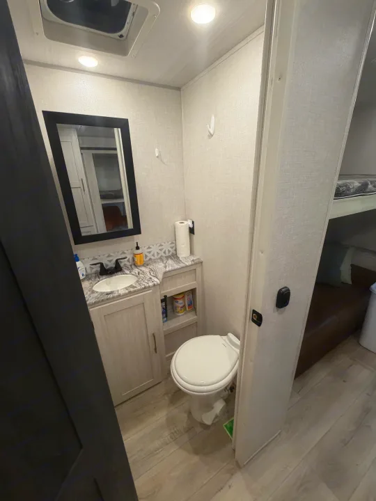 Compact RV rental bathroom, showing a vanity with sink, mirror, toilet, and small storage cubbies.. Forest River Surveyor Legend 2024