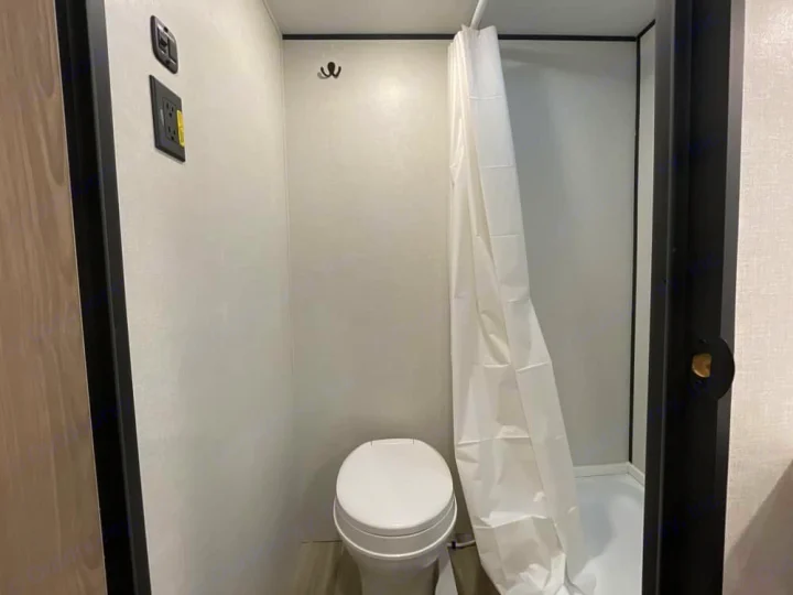 Step into this RV rental's compact wet bath, featuring a toilet, white shower curtain, and wall-mounted controls.. Keystone RV Coleman 13B 2026