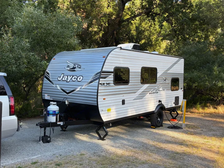 This Jayco travel trailer is an RV rental parked at a shaded campsite among trees, ready for your next adventure.. Jayco Jay Flight SLX 2026