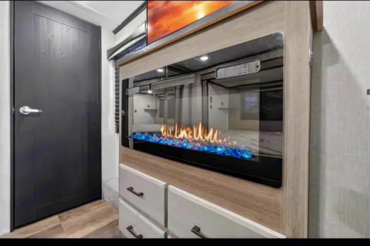 Luxurious RV rental: cozy electric fireplace with blue glass, integrated TV, and stylish storage cabinetry beside a dark door.. Crossroads RV Cameo 2022