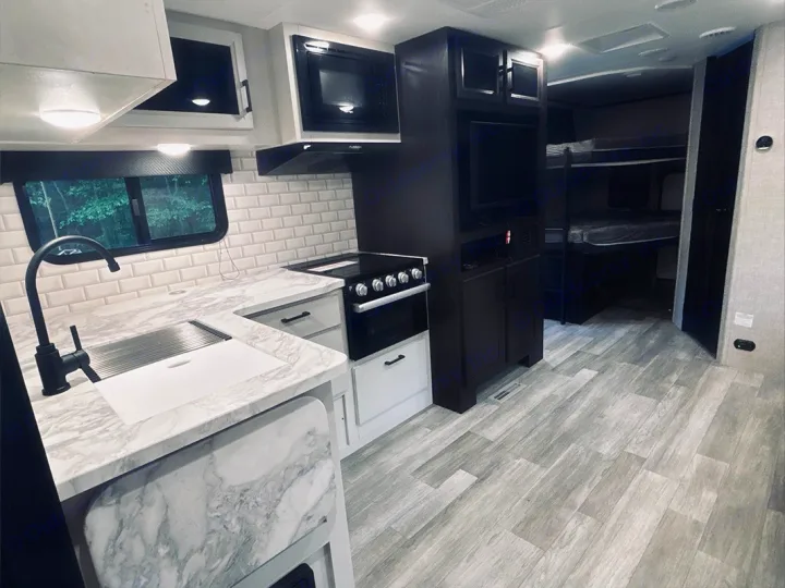 Modern rv rental interior: kitchen with subway tile, marble-look counters, stove, microwave, and bunk beds. Light wood floors.. Jayco Jay Feather 2022