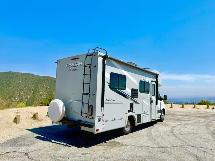 RV rental, light gray, parked at a scenic overlook. Green hills to the left, hazy city view to the right under a clear blue sky.. Coachmen RV Prism Select 24CB 2021