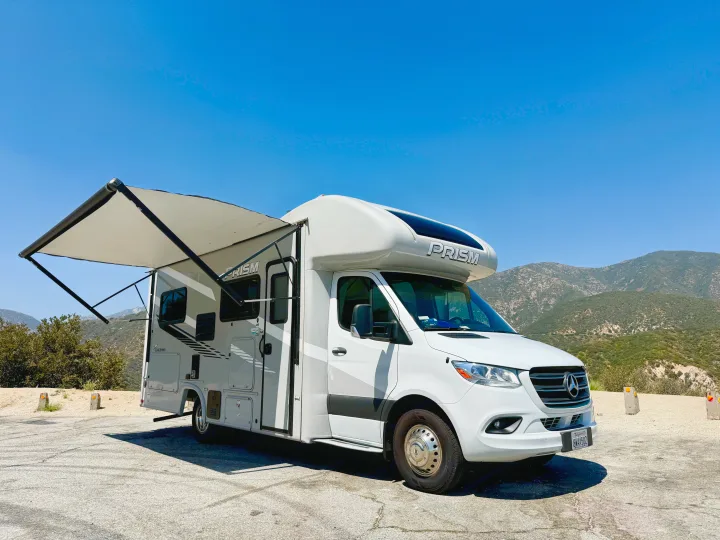 White Prism Coachmen RV rental with extended awning, parked on a paved overlook with green mountains in the background under a blue sky.. Coachmen RV Prism Select 24CB 2021