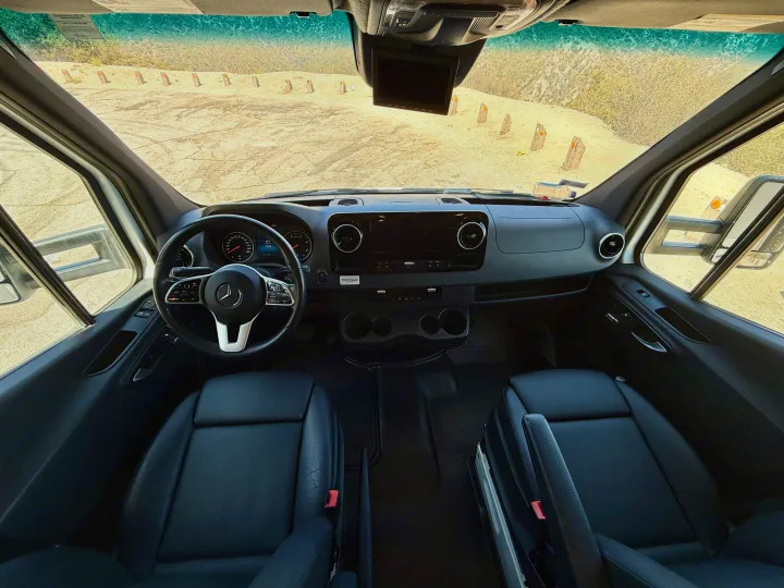 Modern rv rental cockpit interior: black leather seats, Mercedes-Benz steering wheel, and a dusty landscape through the windshield.. Coachmen RV Prism Select 24CB 2021