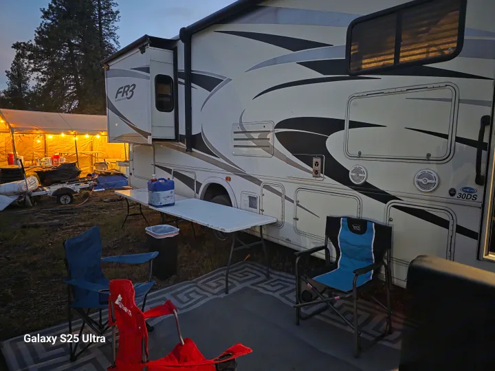 A large RV is parked at a campsite, equipped with chairs, a table, and a brightly lit tent. A comfortable RV rental setup.. Forest River FR3 30DS 2017