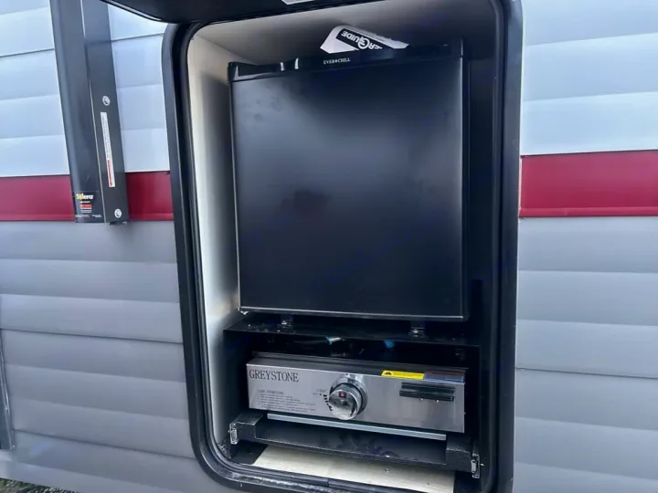 An RV rental's outdoor kitchen features a compact black refrigerator over a GREYSTONE gas cooktop, ideal for outdoor cooking.. Forest River Campsite Reserve 2026