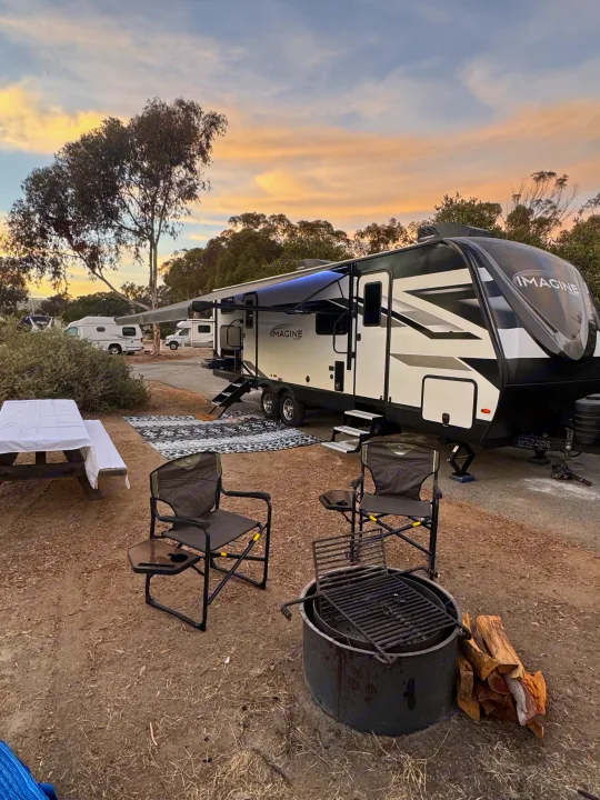 Beautiful sunset at an RV rental campsite featuring a modern travel trailer, fire pit, chairs, and picnic table.. Grand Design Imagine 2025