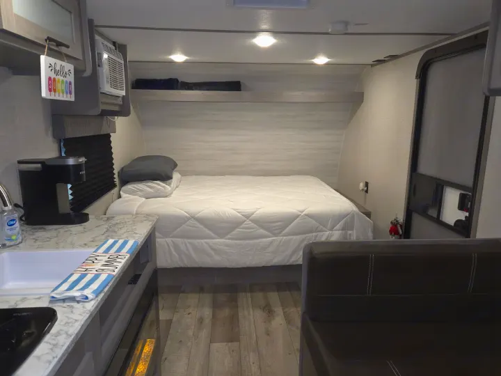 Cozy RV rental interior featuring a comfortable bed with white bedding, a compact kitchen with a sink, and a seating area.. Keystone RV CM17R23 2023