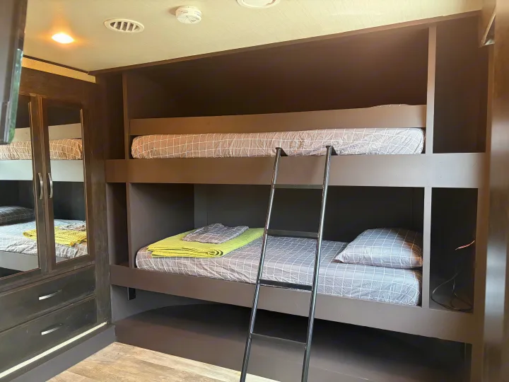 Cozy RV rental bunk beds with grey bedding, a green blanket, and a ladder. Ideal for compact sleeping in a travel trailer.. Grand Design Momentum 2017
