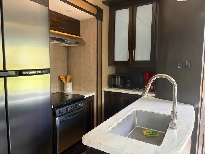 Modern RV rental kitchen with stainless steel fridge, stove, sink, microwave, and coffee maker. Dark wood cabinets and light countertops.. Grand Design Momentum 2017
