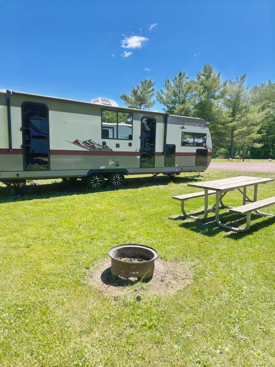 An RV rental parked at a serene campsite, complete with a picnic table and fire pit under a clear blue sky.. Forest River Campsite Ultra 26CJL 2025