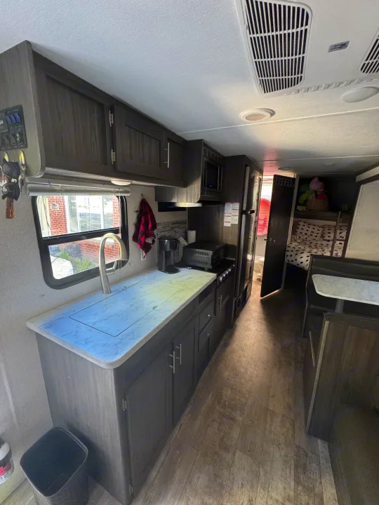 Inside a modern rv rental, featuring a full kitchen with sink, stove, fridge, and a dining area. Bunk beds seen beyond.. Forest River Shasta 2021