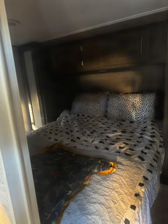 Inside a cozy rv rental: a neatly made bed with spotted pillows, a black-patterned duvet, and a dinosaur blanket under dark wood cabinets.. Forest River Shasta 2021