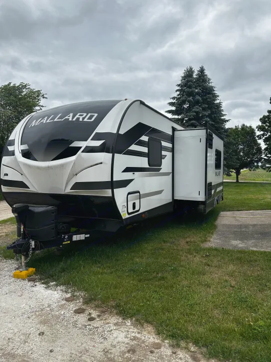 White and black Mallard RV rental, a travel trailer with an extended slide-out, parked on grass beneath an overcast sky.. Heartland RVs Mallard 2022