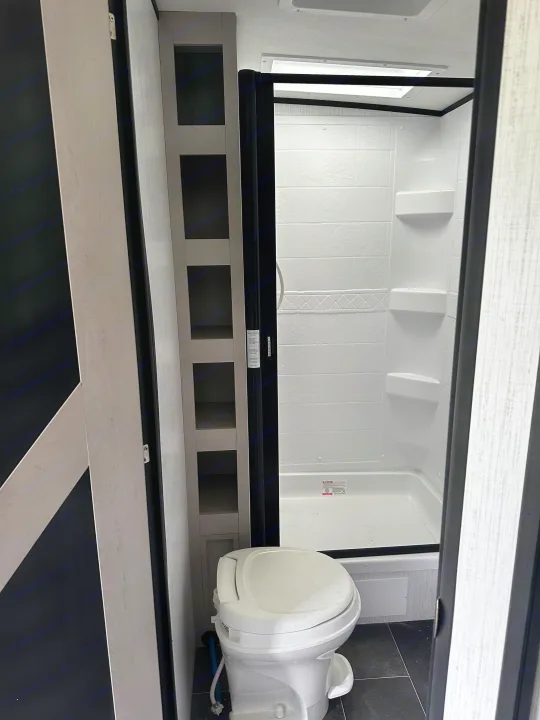 Compact RV rental bathroom featuring a white toilet, enclosed shower stall, and tall storage shelves.. Heartland RVs Mallard 2022