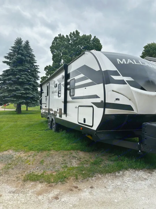 This white Mallard travel trailer, perfect for rv rental, is parked on grass with tall trees & a cloudy sky for your next adventure.. Heartland RVs Mallard 2022