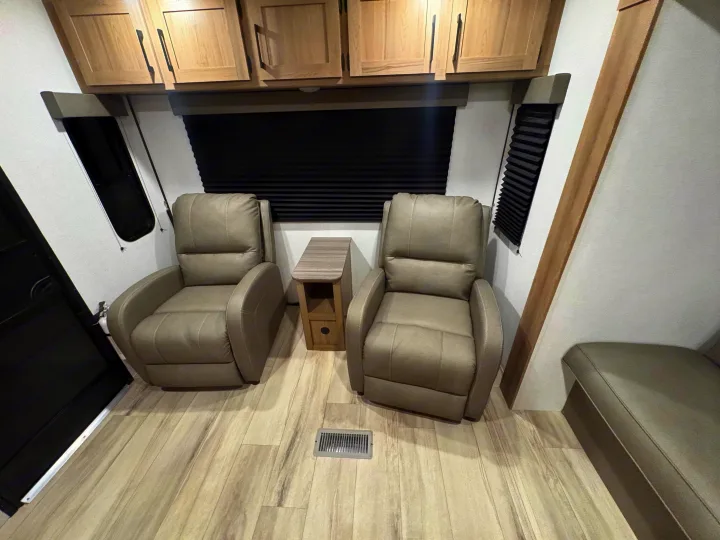 RV rental interior with two tan recliner chairs, a side table, overhead cabinets, wood-look flooring, and dark window blinds.. Keystone RV Other 2026