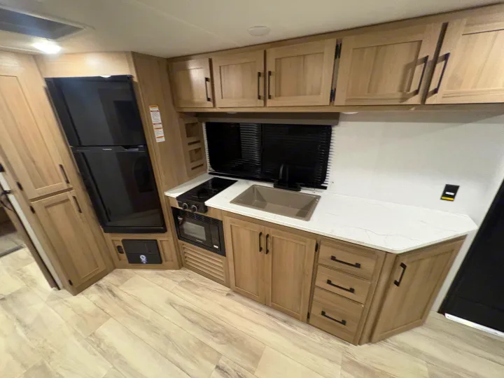 Inside an RV rental, a compact kitchen features a black fridge, stove, microwave, sink, and light wood cabinets with white counters.. Keystone RV Other 2026