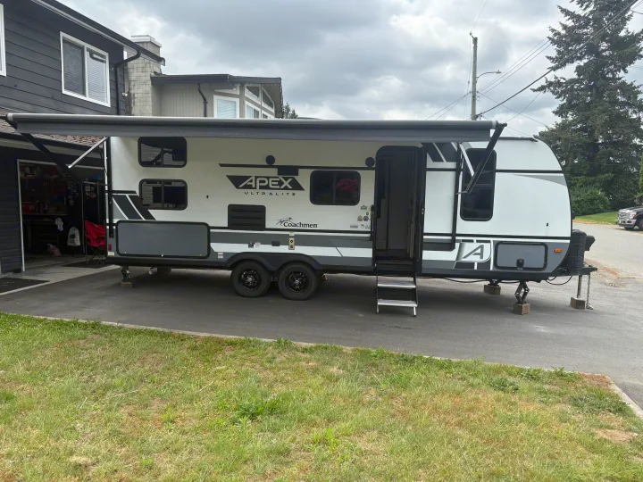 White and gray Apex Ultralite RV rental parked on a driveway next to a house with its awning extended.. Coachmen Apex 2022