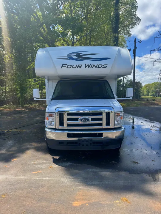 Front view of a white Ford Four Winds Class C RV rental parked on a wet asphalt lot with trees in the background.. Thor Four Winds 2023