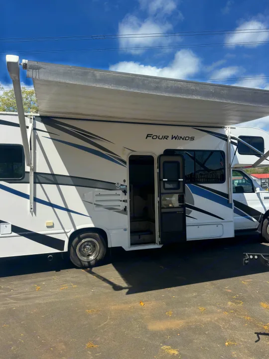White Four Winds RV rental with extended awning & open entrance, parked on asphalt under a blue sky.. Thor Four Winds 2023