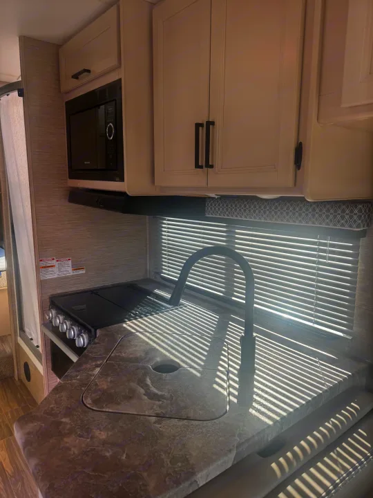 RV rental kitchen interior with microwave, range, sink, and cabinets. Sunlight streaks across the counter.. Thor Four Winds 2023