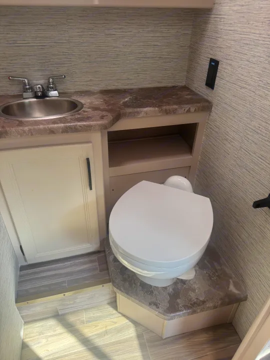 This RV rental features a functional, clean bathroom with a compact sink, toilet, shelving, and textured wall coverings.. Thor Four Winds 2023