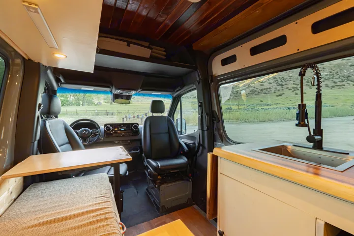 RV rental van interior with front seats, a table, and a kitchenette with sink, offering a scenic view of hills and grassland.. MERCEDES-BENZ SPRINTER 2500 170 WB 2022