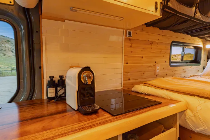 RV rental van interior featuring a sleek coffee maker, induction cooktop, wooden counter, and cozy bed setup.. MERCEDES-BENZ SPRINTER 2500 170 WB 2022