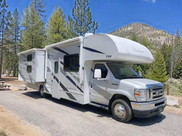 Explore nature with this Class C rv rental. Silver motorhome with slide-out parked on gravel, surrounded by trees & mountains.. Thor Freedom Elite 2022