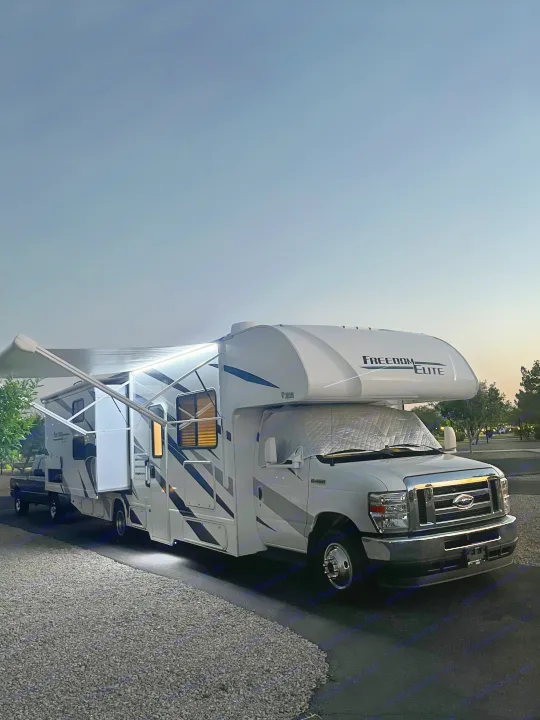 White Freedom Elite rv rental with an extended lighted awning, towing a truck, parked at dusk. Perfect for travel.. Thor Freedom Elite 2022