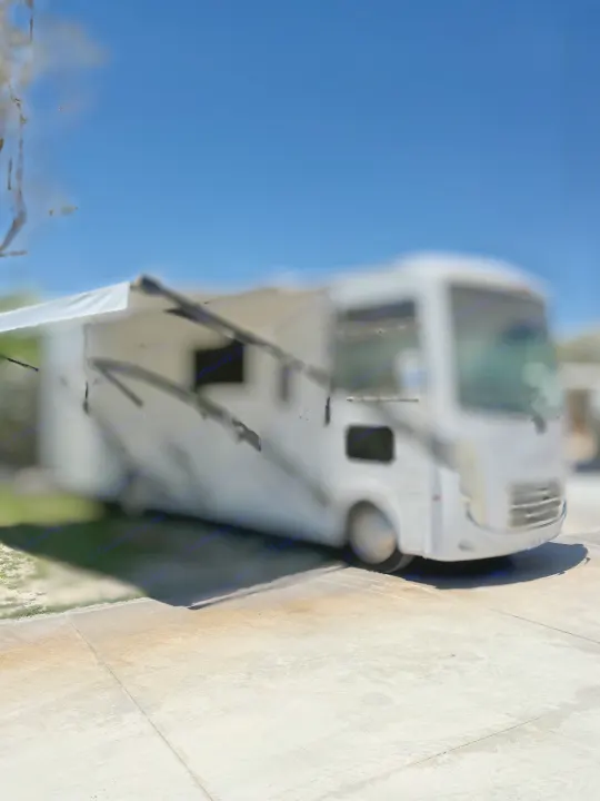 Blurred white RV rental with awning extended, parked on concrete under a blue sky. Perfect for your next adventure!. Thor Hurricane 2024