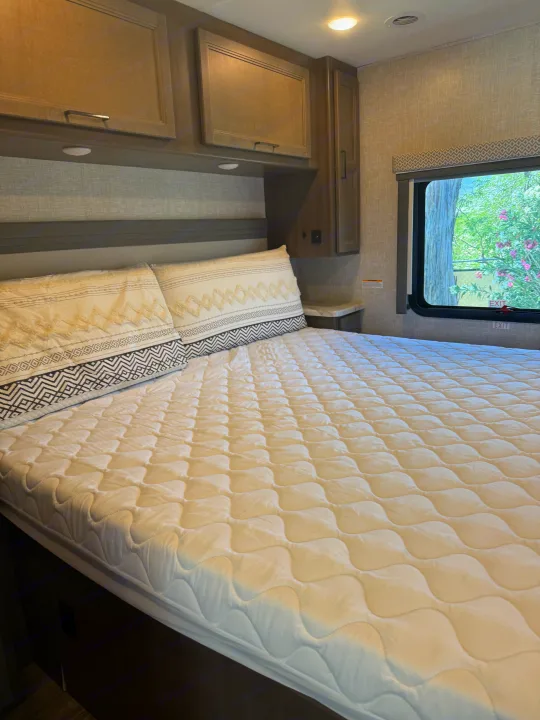 Cozy RV rental bedroom featuring a quilted bed with decorative pillows, overhead cabinets, and a bright window with a nature view.. Thor Hurricane 2024
