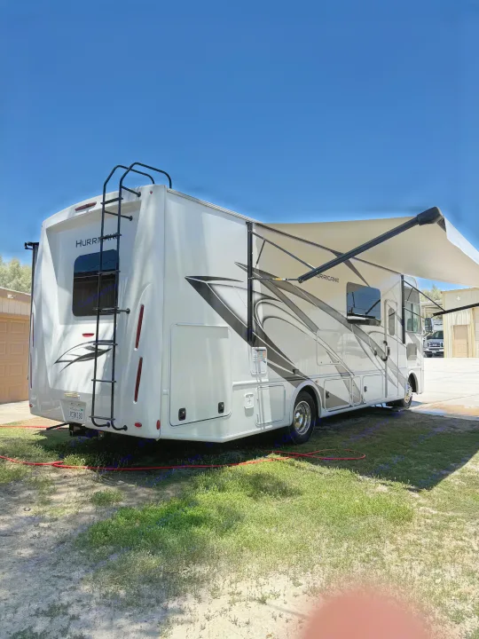 White Hurricane RV rental motorhome with its awning extended, parked on grass under a clear blue sky.. Thor Hurricane 2024