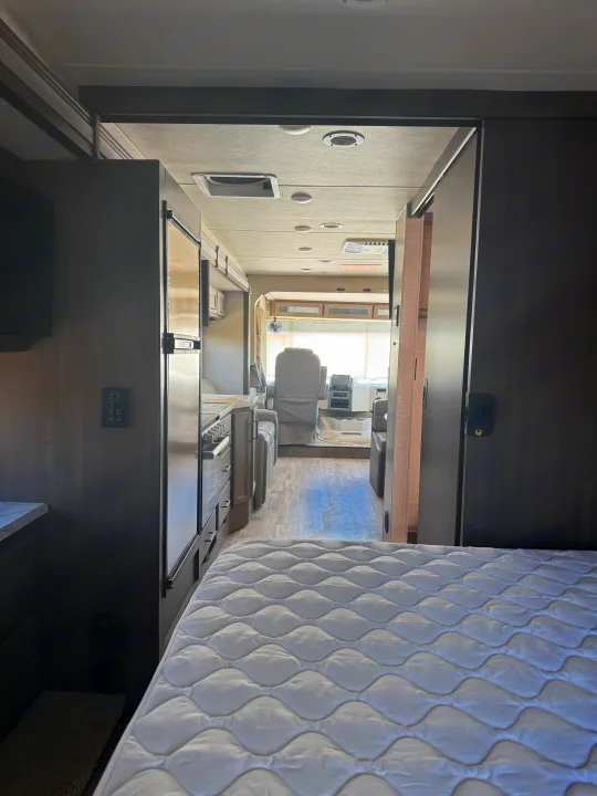 View from an RV rental bedroom showing a plush mattress, leading to a kitchen, and the driver's cockpit with seats and dashboard.. Thor Hurricane 2024