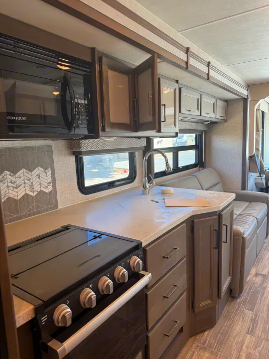 Inside a clean RV rental: kitchen area with microwave, stovetop, sink, and cabinets, plus a cozy sofa for relaxing.. Thor Hurricane 2024