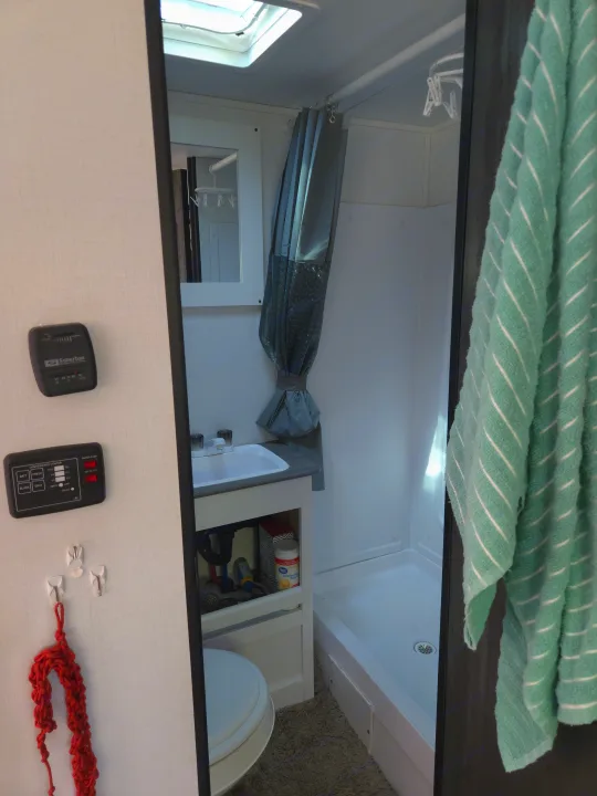 RV rental bathroom with a white sink, toilet, compact shower stall, and wall panels for thermostat & tank levels. A green towel hangs right.. Keystone RV Hideout 2022