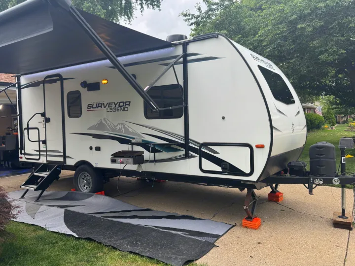 A white Surveyor Legend travel trailer, with an extended awning, outdoor grill, and mat, setup for an RV rental.. Forest River Surveyor Legend 2021