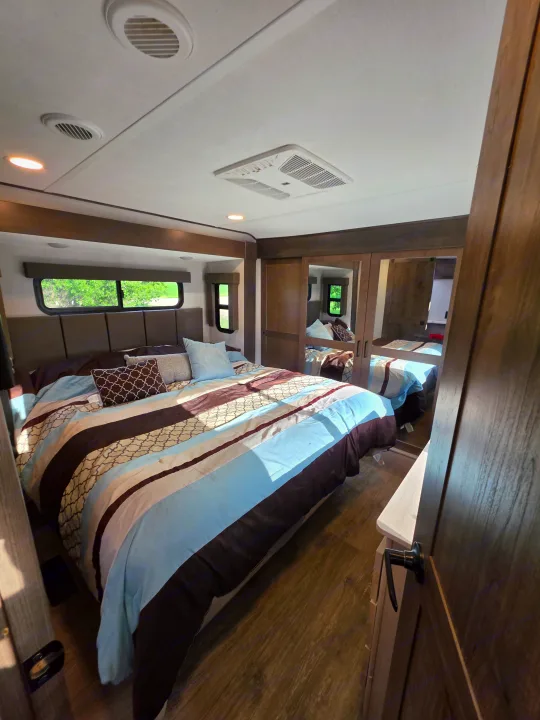 RV rental bedroom featuring a striped bed, windows overlooking greenery, mirrored closet, and ceiling AC/vents.. Alliance RV Valor 40V13 2021