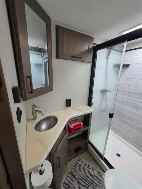 Modern RV rental bathroom featuring a sink, mirror, toilet, and tiled shower, with vanity storage and a first aid kit.. Alliance RV Valor 40V13 2021