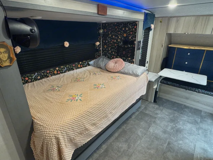 This RV rental interior features a cozy bed with a textured blanket, floral wallpaper, a fan, and a compact dinette area.. Forest River Salem FSX 2025