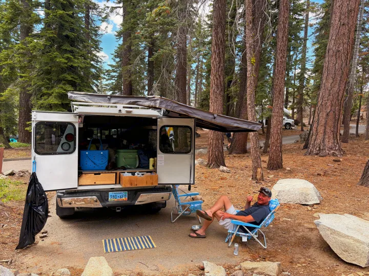 Man relaxing at a wooded campsite next to his open truck camper, ready for adventure. Explore nature with an RV rental!. Ford Ford F150 2017