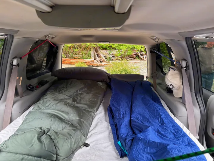 Inside a car camping setup: two sleeping bags & pillows for an RV rental road trip. Trees and logs visible outside.. chevy uplander 2006