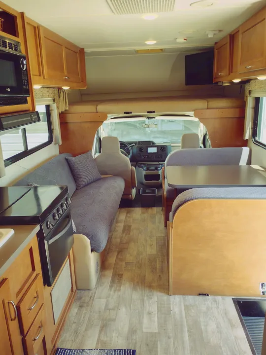 Inside a clean RV rental: kitchen counter with stove, dinette, and driver's cab. Features wood cabinets & light flooring.. Winnebago Minnie Winnie 2017