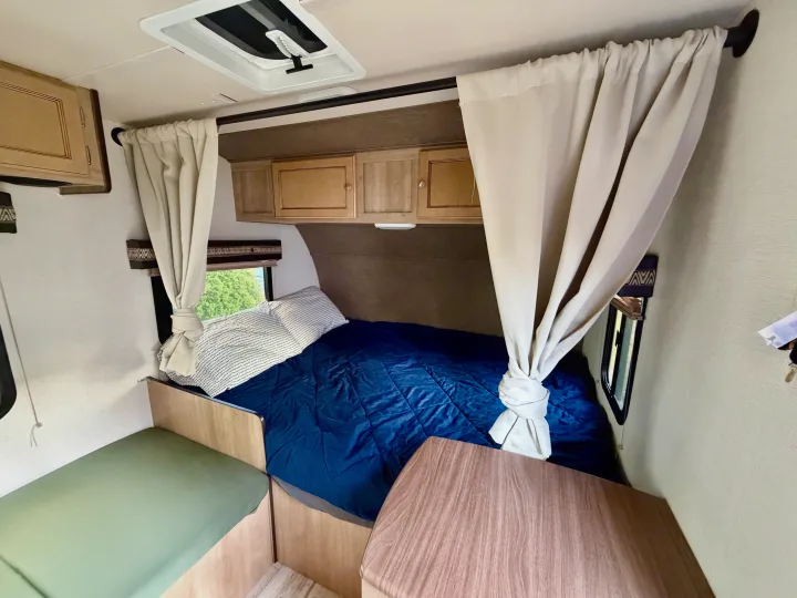 Inside this RV rental: a snug sleeping area with blue bedding, tied curtains, overhead cabinets, and a side dinette.. Palomino Real Lite Mini 2017